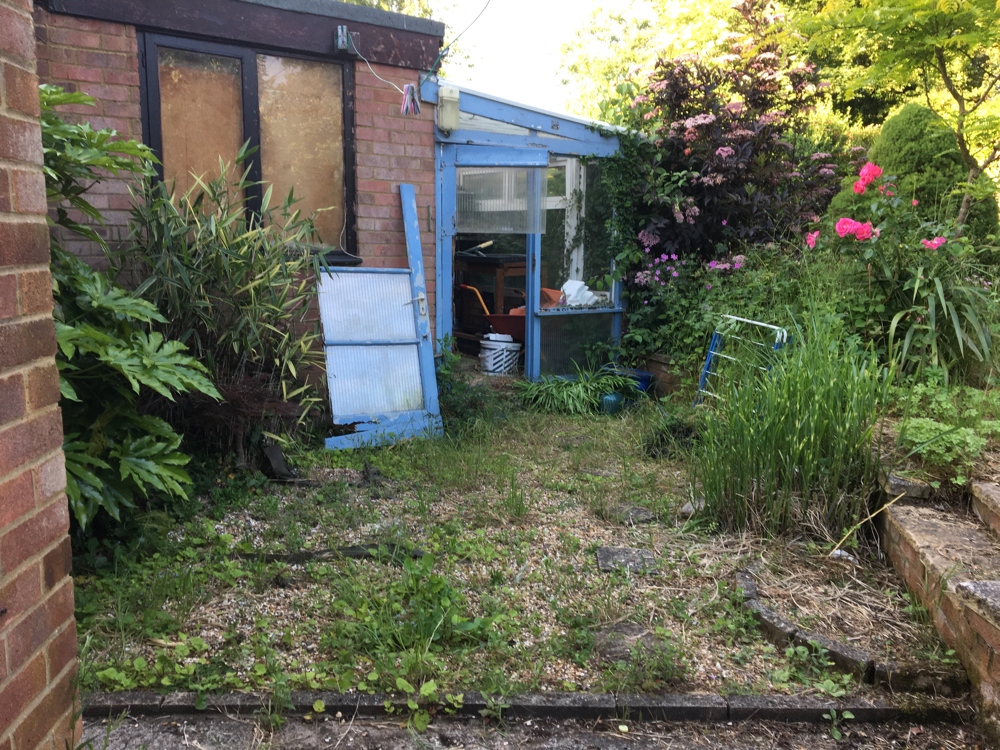 Greenhouse with. broken door and a very weedy gravel section in fron of it.